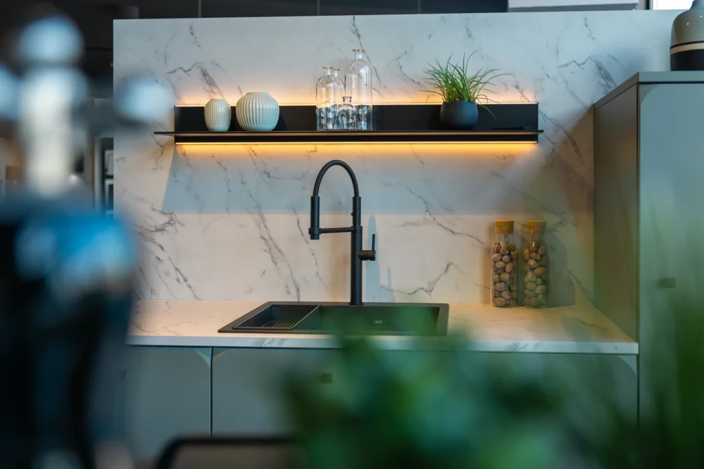 Schwarze Spüle mit Profi-Armatur vor Marmor-Rückwand und beleuchtetem Wandboard